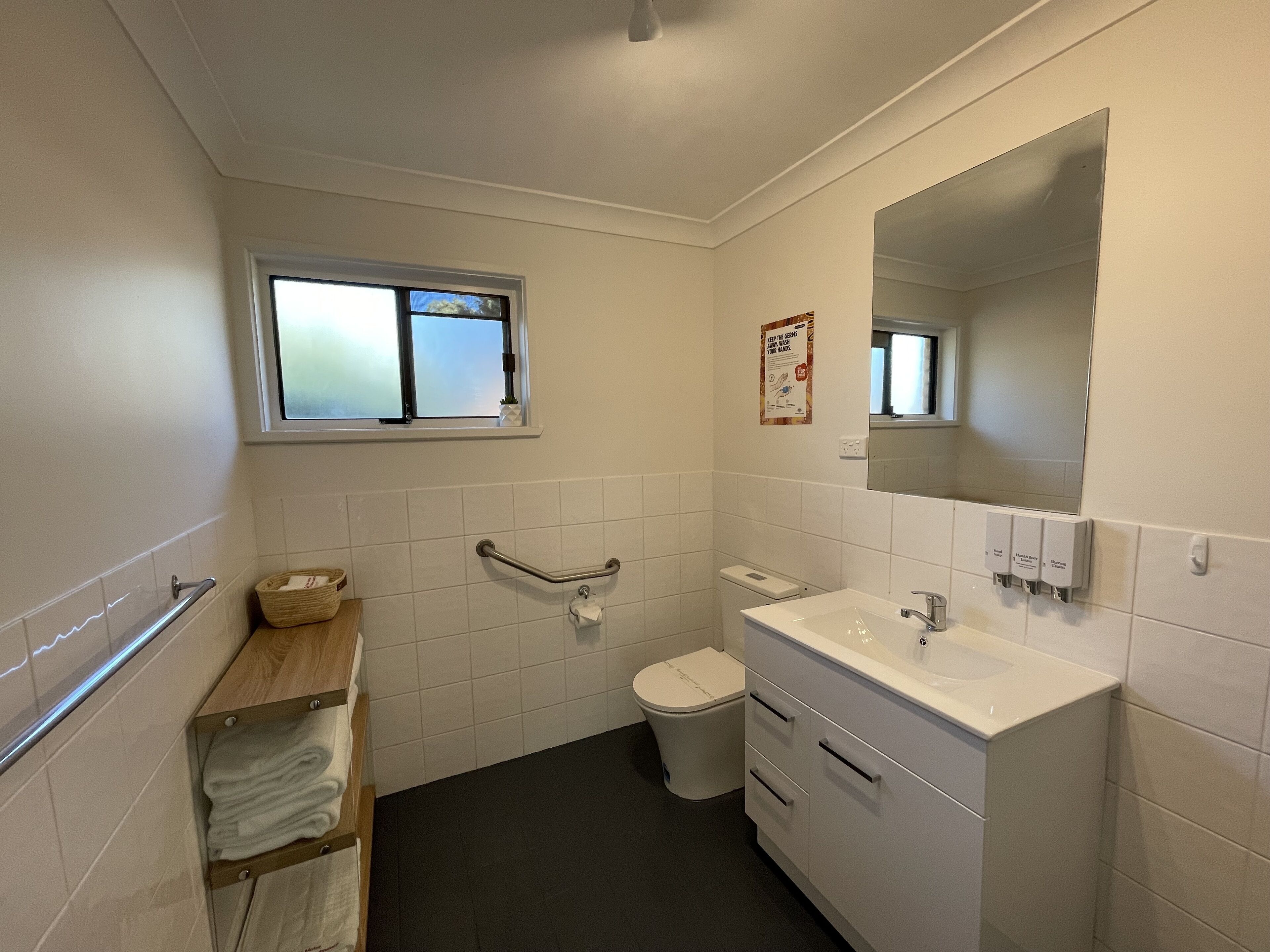 Ein kleines, sauberes Badezimmer mit weißen Fliesen, Toilette, Waschbecken, Spiegel und Fenster.