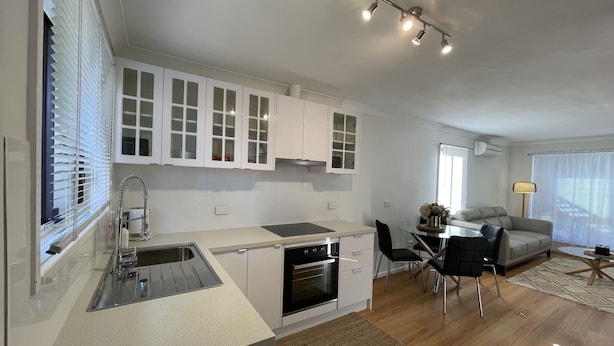 Una cocina moderna con armarios blancos, horno empotrado y comedor con mesa y sillas.