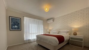 Un dormitorio con cama, mesita de noche, lámpara, aire acondicionado instalado en la pared y ventana con cortinas translúcidas.