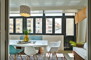 A dining area with a table, chairs, a vase with flowers, and a view of buildings through large windows.