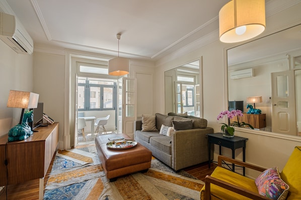 A well-lit living room with a sofa, ottoman, and a coffee table. There is a dining area with chairs and a table, and a large mirror on the wall.