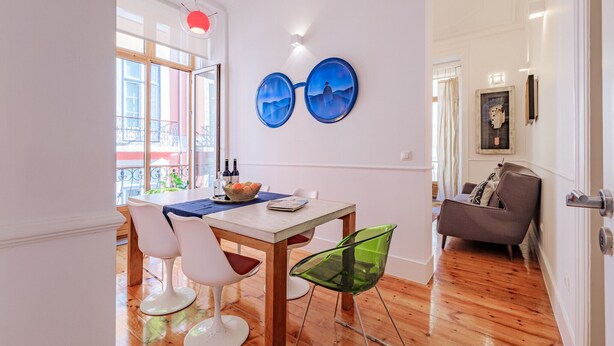 A dining area with a wooden table, white chairs, and a green chair. There is a window with a view of a balcony. - Central 3BR/2BR Apartment | Balconies| Elevator| Baixa (Lisboa ciudad)
