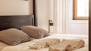A neatly made bed with patterned pillows, a bedside lamp, and a window with curtains. - Central 3BR/2BR Apartment | Balconies| Elevator| Baixa (Lisboa ciudad)