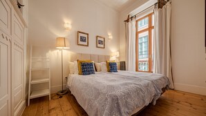 A bedroom with a large bed, wooden flooring, a wardrobe, a floor lamp, and a window with curtains. - Central Apartment 2BR/2BA | Balcony | Lift in Baixa (Lisboa ciudad)