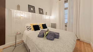 Ein ordentlich bezogenes Bett mit einer gemusterten Tagesdecke, Kissen und gefalteten Handtüchern. Neben dem Bett steht ein kleiner Tisch mit einer Lampe und einem Buch.