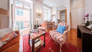 A spacious living room with a red carpet, a wooden coffee table, striped armchairs, and a dining area with a table and chairs. - Central 2BR/2BA Apartment Balcony Elevator in Baixa (Lisboa ciudad)