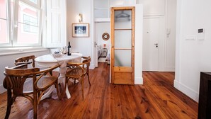 A room with a wooden floor, white walls, a round table with chairs, a door with a frosted glass panel, and a window with a view of buildings. - Central 2BR/2BA Apartment Balcony Elevator in Baixa (Lisboa ciudad)