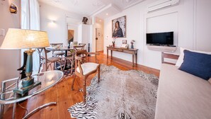 A modern hotel room with a wooden floor, a zebra-patterned rug, a bed with a blue pillow, a TV, a desk with a lamp, and a dining area with chairs and a table. - Baixa Apartment 2BR|2BA with Balcony, Lift (Lisboa ciudad)