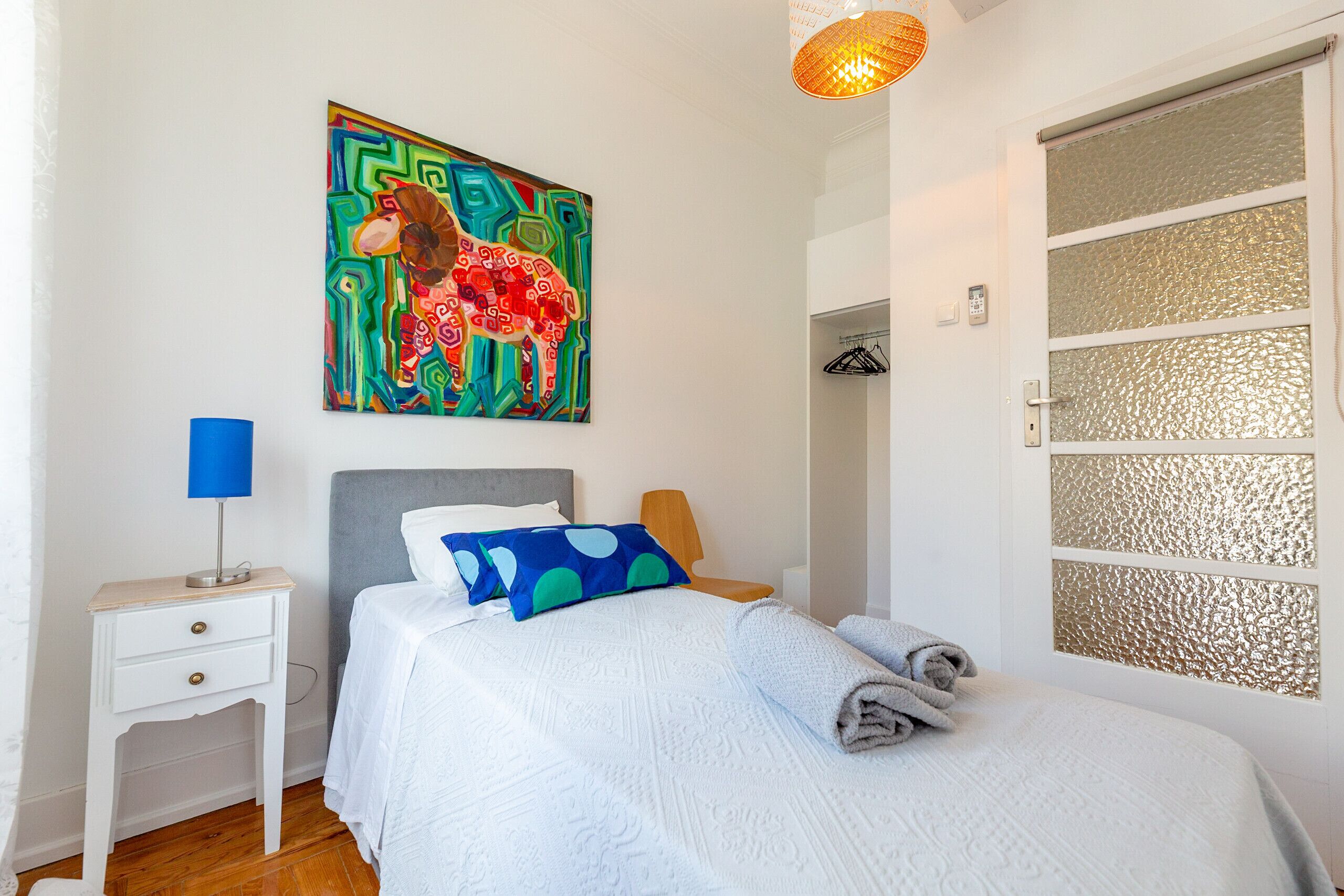 Een net opgemaakt bed met een witte dekbedovertrek en blauwe kussens, een klein nachtkastje met een lamp, en een kleurrijk abstract schilderij aan de muur.