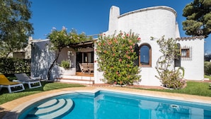 A white villa with a swimming pool, a patio with chairs, and a garden with blooming flowers.