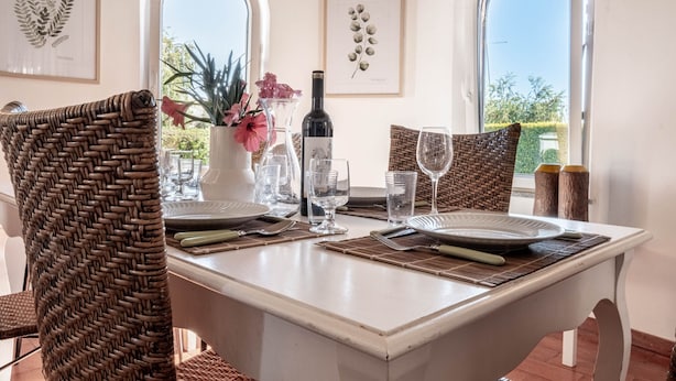 A dining table set with plates, cutlery, and glasses, surrounded by wicker chairs, with a wine bottle and flowers on the table.