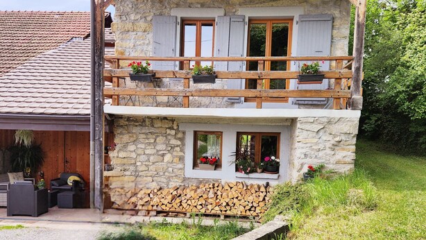 A two-story stone house with a wooden balcony, a small attached garage, and a garden area with potted plants.