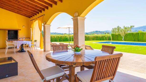 An outdoor patio with a round wooden table and chairs, a TV, and a view of a swimming pool and greenery. - ES MUSSOL - Villa with private pool in SA POBLA. Free WiFi (Sa Pobla)