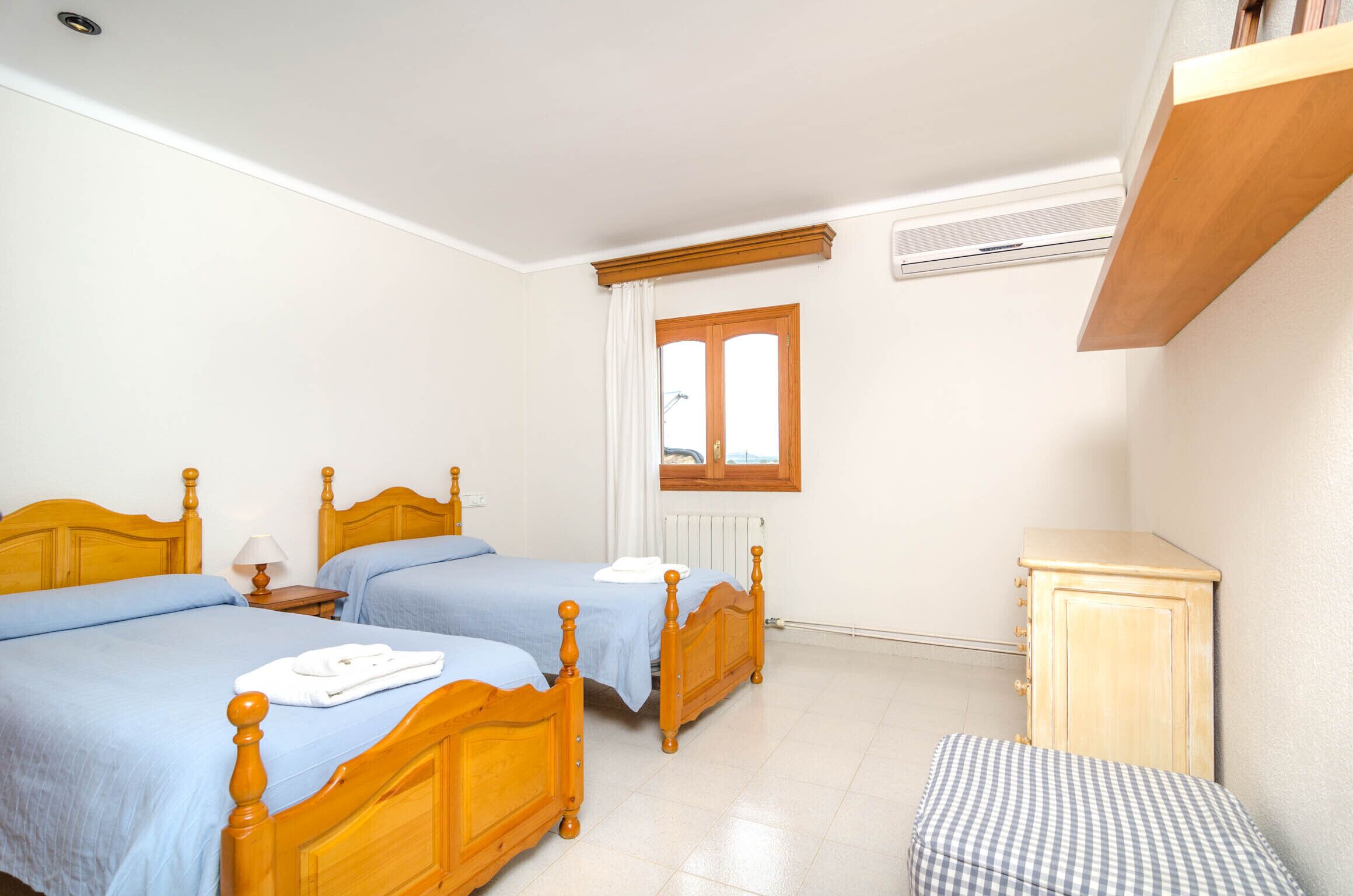 A bedroom with two wooden beds, a small bedside table with a lamp, a radiator, a window with curtains, and a wooden cabinet.