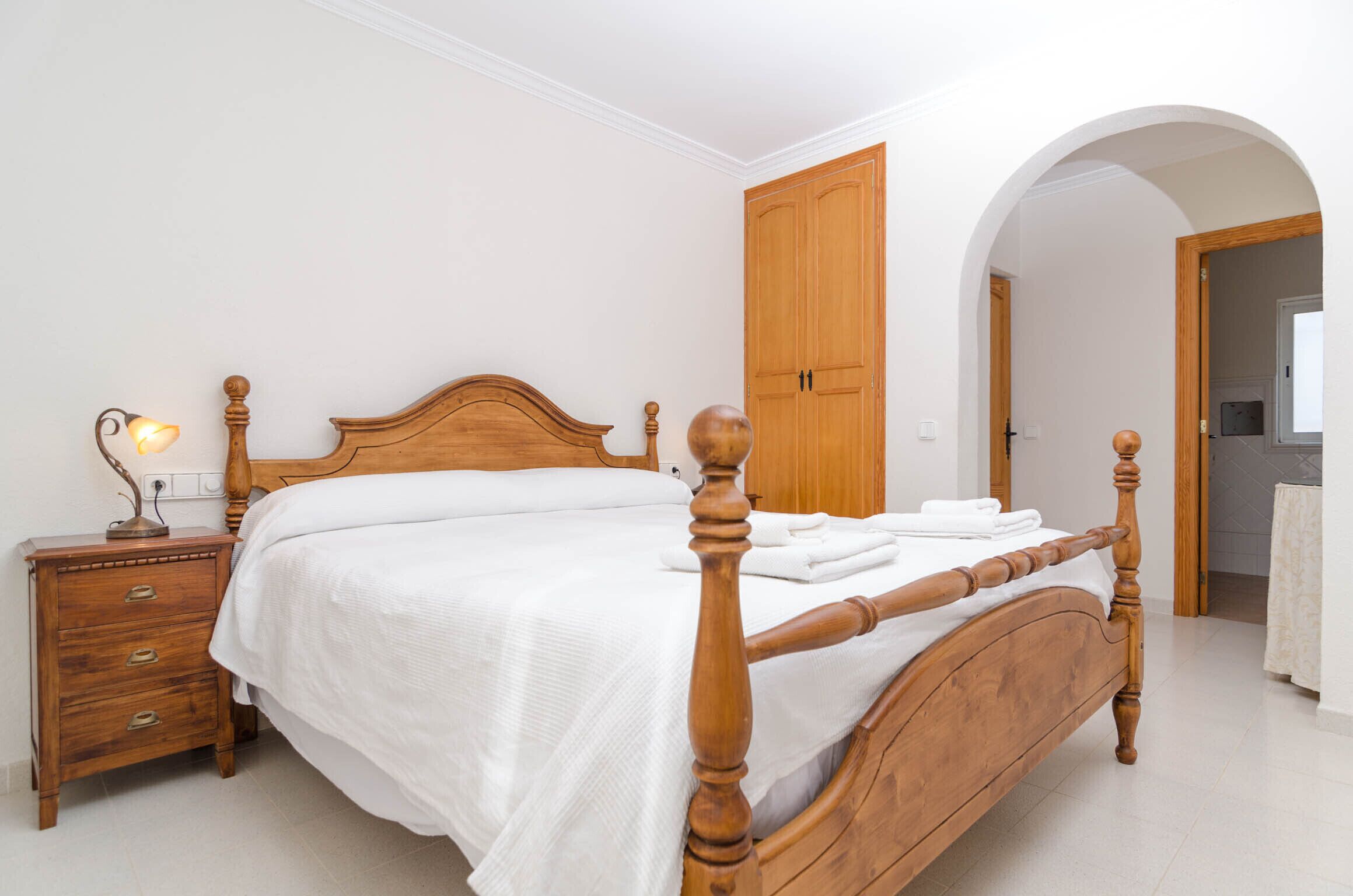 Uma cama bem arrumada com cabeceira de madeira, uma mesa de cabeceira com abajur e um guarda-roupa num quarto com paredes brancas e uma porta em arco.