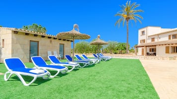 Een rij blauwe ligstoelen met rieten parasols in een binnenplaats met een palmboom en een gebouw met een betegeld dak.
