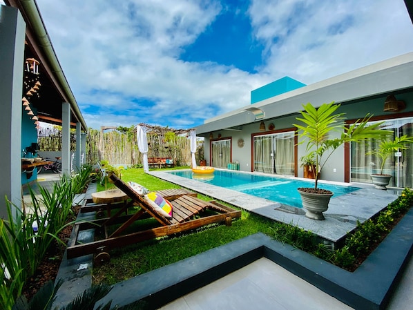 Un área de piscina moderna al aire libre con terraza de madera, sillones de descanso y un pequeño jardín con plantas en macetas.