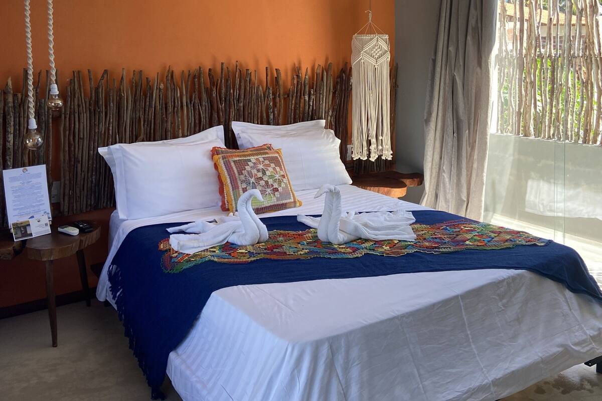 A hotel room with a large bed, a wooden headboard, a hanging light fixture, and a view of palm trees through the window.