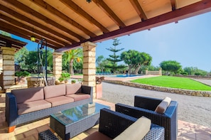 Une terrasse extérieure avec des meubles en osier, un pilier en pierre et une vue sur une piscine et de la verdure.