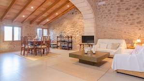 Un vaste espace de vie agrémenté d’un mur en pierre, de poutres apparentes en bois au plafond, d’une table à manger avec des chaises, d’un canapé et d’un téléviseur.