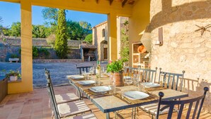 Un espace extérieur aménagé pour les repas, avec un mur en pierre, une pergola en bois et, en arrière-plan, une vue sur un bâtiment en pierre.
