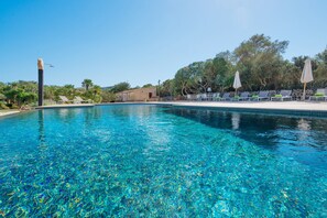 Una piscina azul cristalina con sillas de descanso y sombrillas, rodeada de árboles y un muro de piedra.