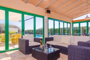 A covered outdoor seating area with wicker furniture and a glass table, overlooking a swimming pool and greenery. - CAN RITO - Villa with private pool in FELANITX. Free WiFi (Felanitx)