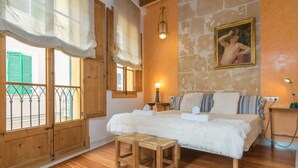 A bedroom with a wooden floor, a bed with white bedding, a small wooden stool, a nightstand with a lamp, and a painting on the wall.