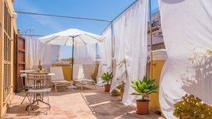 Eine Terrasse mit Tisch, Stühlen und Sonnenschirmen. Es stehen Blumentöpfe auf der Terrasse und es hängt ein weißer Vorhang.