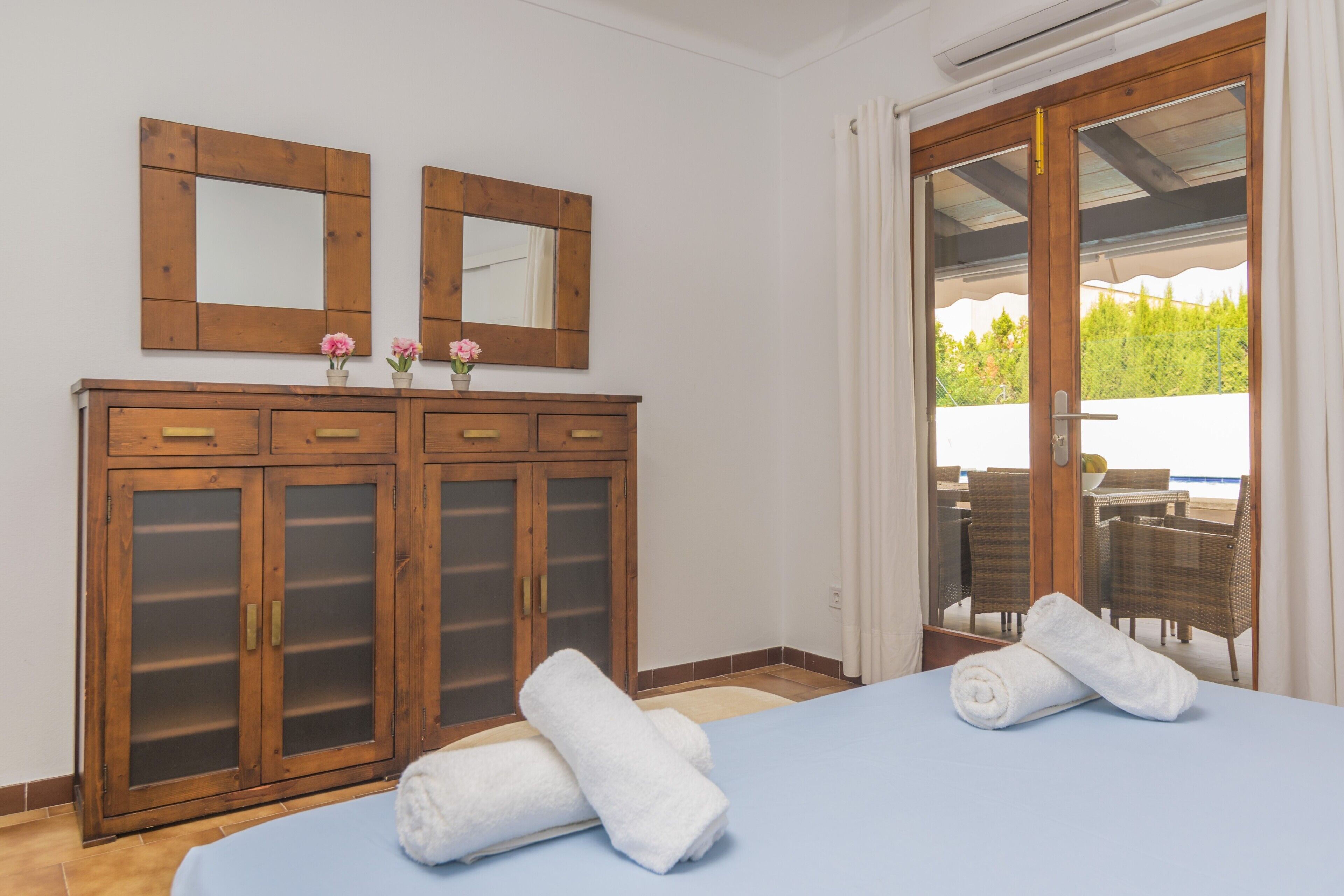 Ein Zimmer mit einem Bett, zwei Nachttische mit Glastüren, einem Spiegel und Blick auf Gartenmöbel durch eine Glastür.