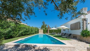 Une piscine rectangulaire entourée d’une terrasse en pierre, elle-même bordée d’une végétation luxuriante et d’un bâtiment blanc à la façade en pierre.