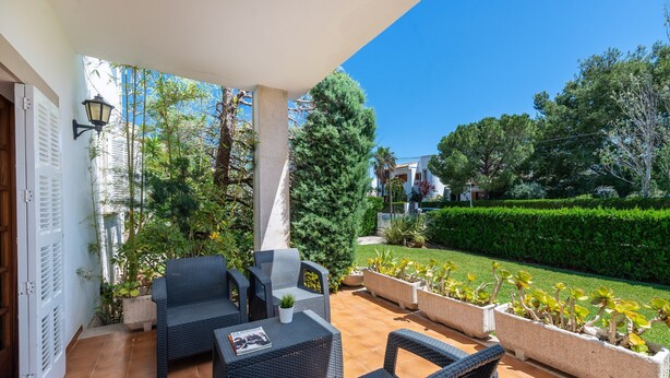 Une terrasse couverte, aménagée avec des meubles en osier, des plantes en pot et offrant une vue sur un jardin bien entretenu et les bâtiments voisins.