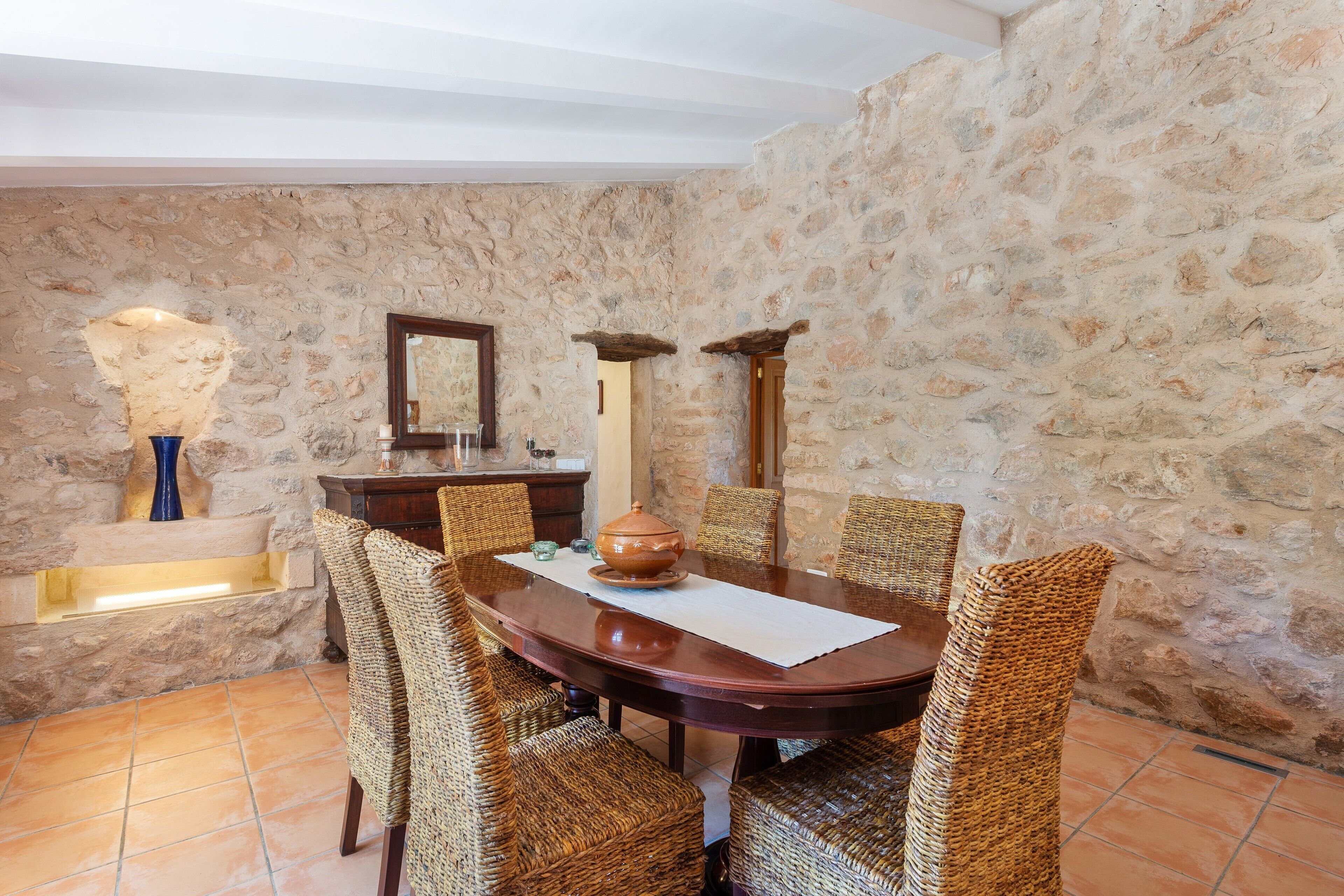 A dining area with a stone wall, a wooden table, wicker chairs, and a large vase.