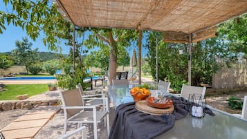 An outdoor patio with a glass table, white chairs, a woven umbrella, and a pool in the background.