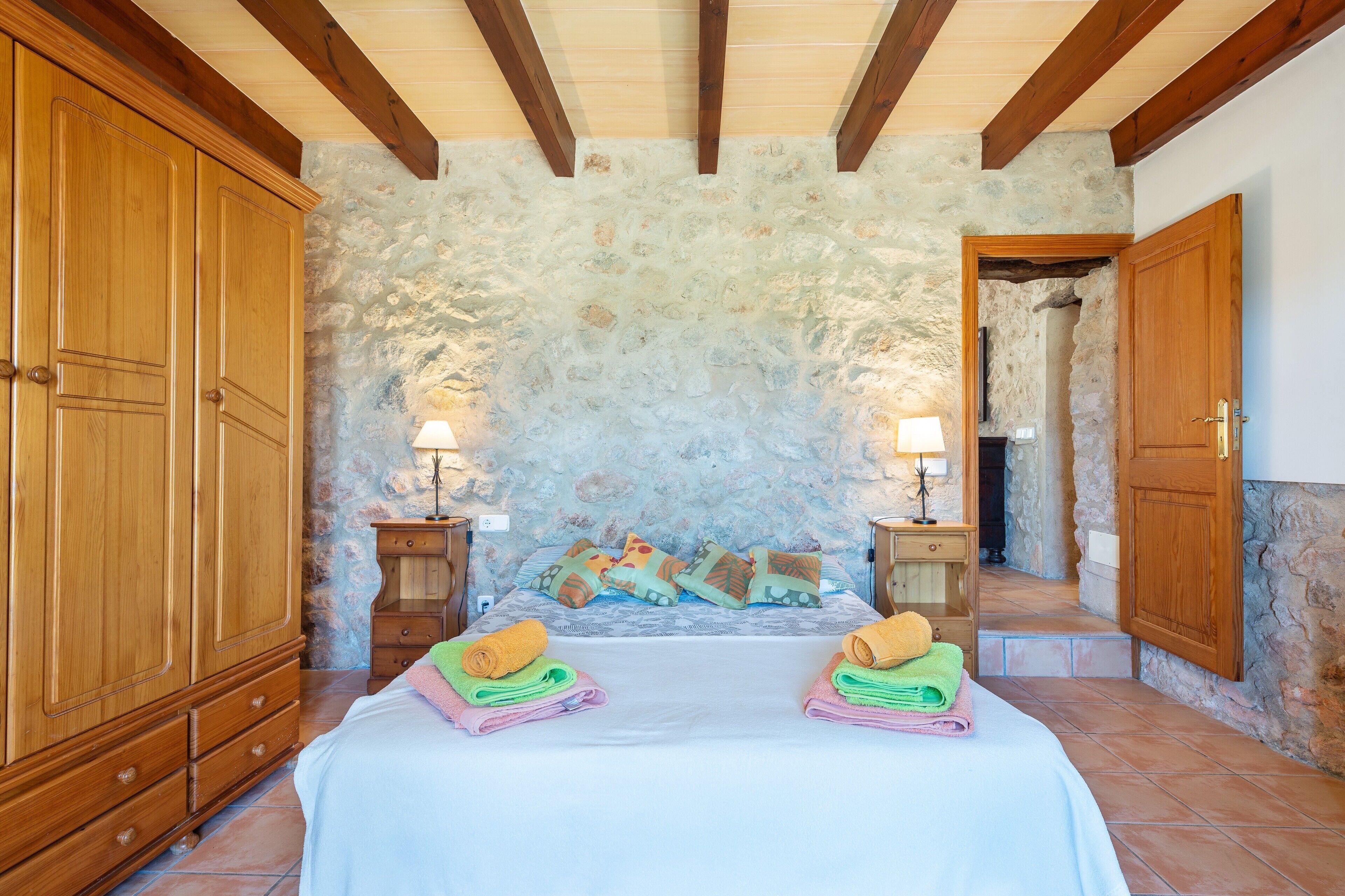 A bedroom with a stone wall, wooden wardrobe, bed with colorful towels, and two bedside tables with lamps.