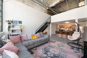 Un espacio habitable moderno con un sofá gris, cojines coloridos, un tapete estampado, una silla colgante, una escalera y una zona de cocina.