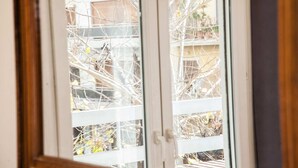 A view through a window with a white frame, displaying a balcony with bare branches and some yellow leaves, and a building in the background.