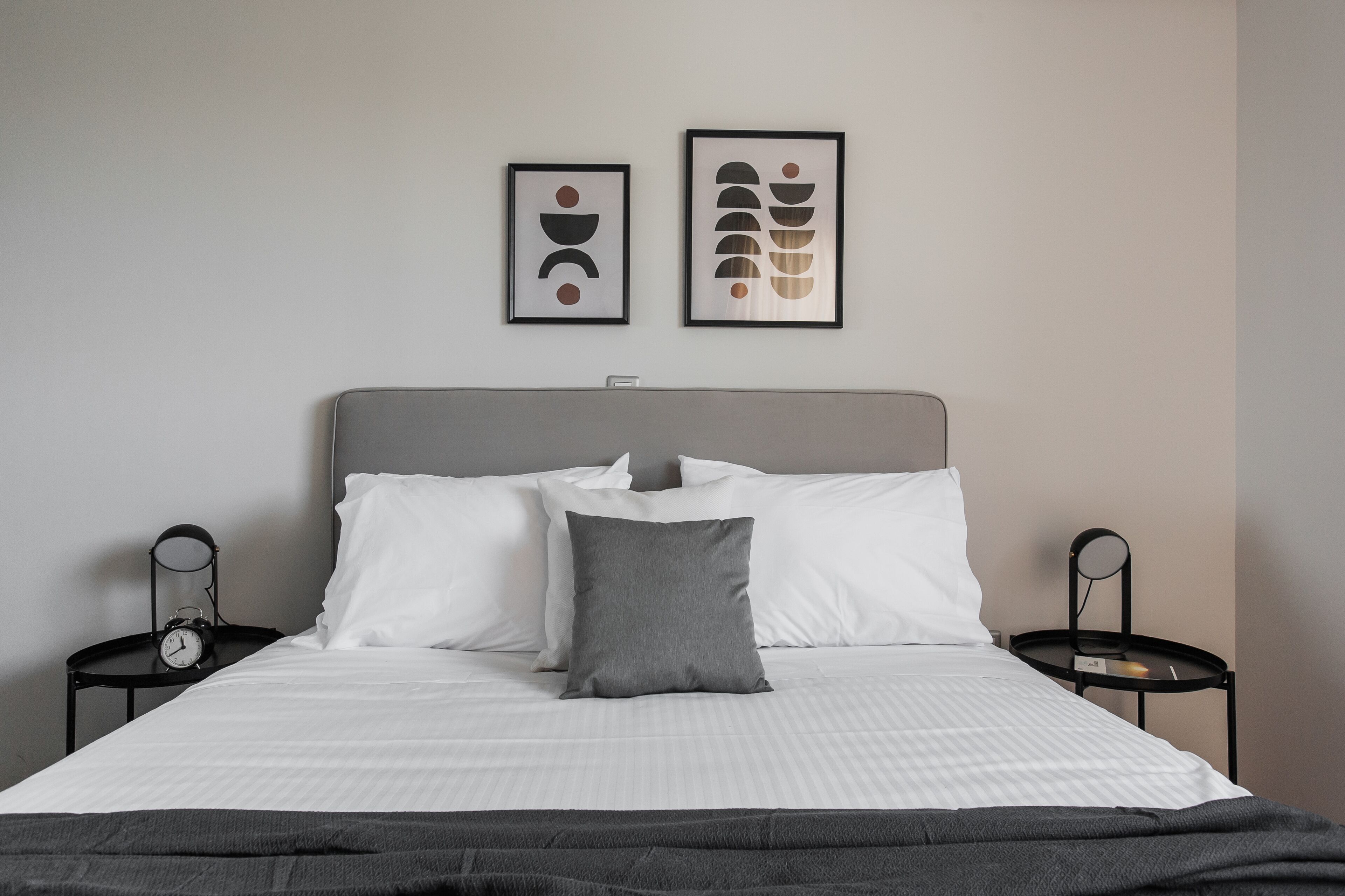 Ein ordentlich bezogenes Bett mit weißer und grauer Bettwäsche, zwei gerahmte abstrakte Kunstwerke an der Wand und ein kleiner Tisch mit einer Uhr auf jeder Seite.
