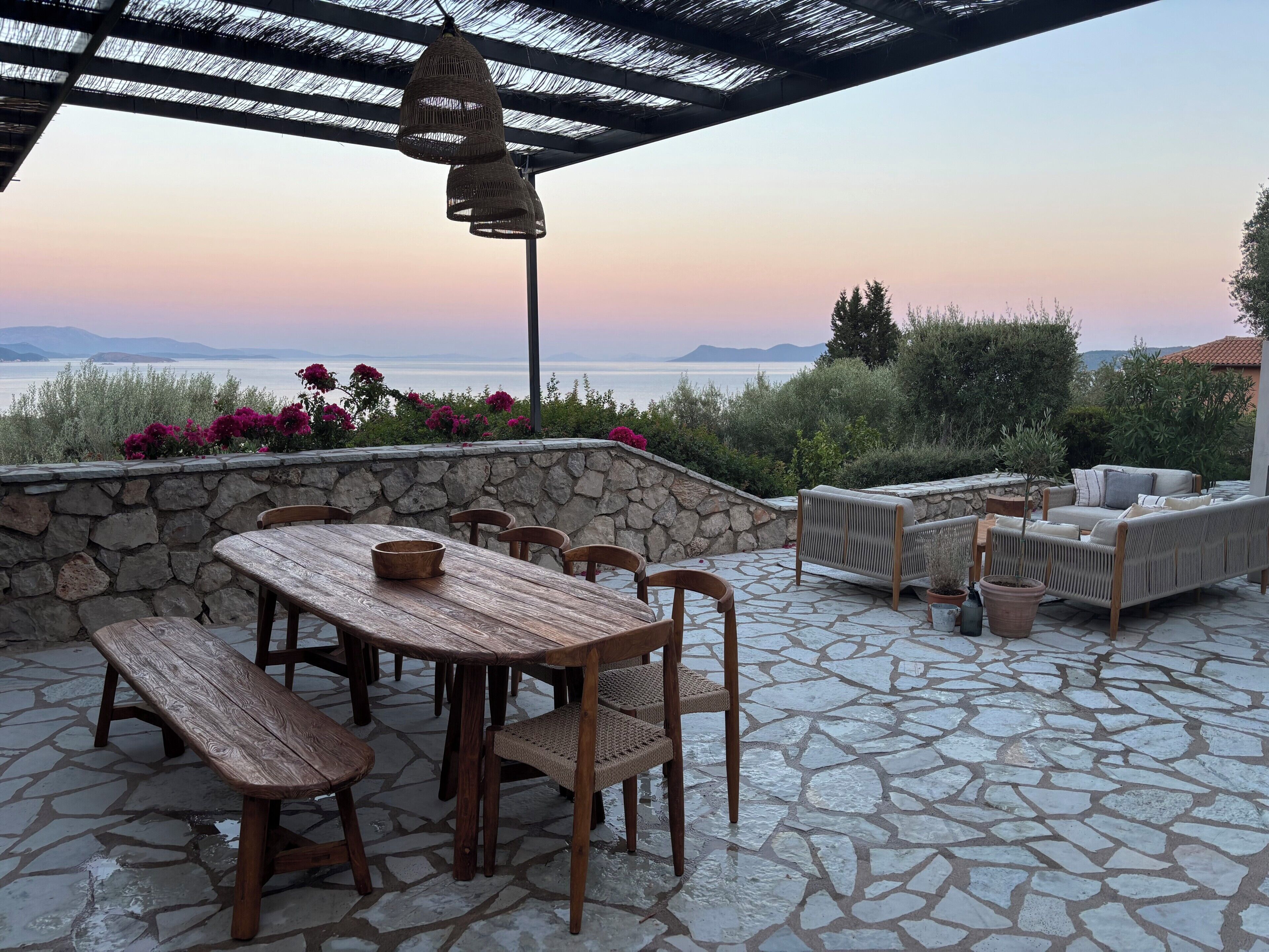 Un patio al aire libre con suelo de piedra, mobiliario de madera y vista al mar.