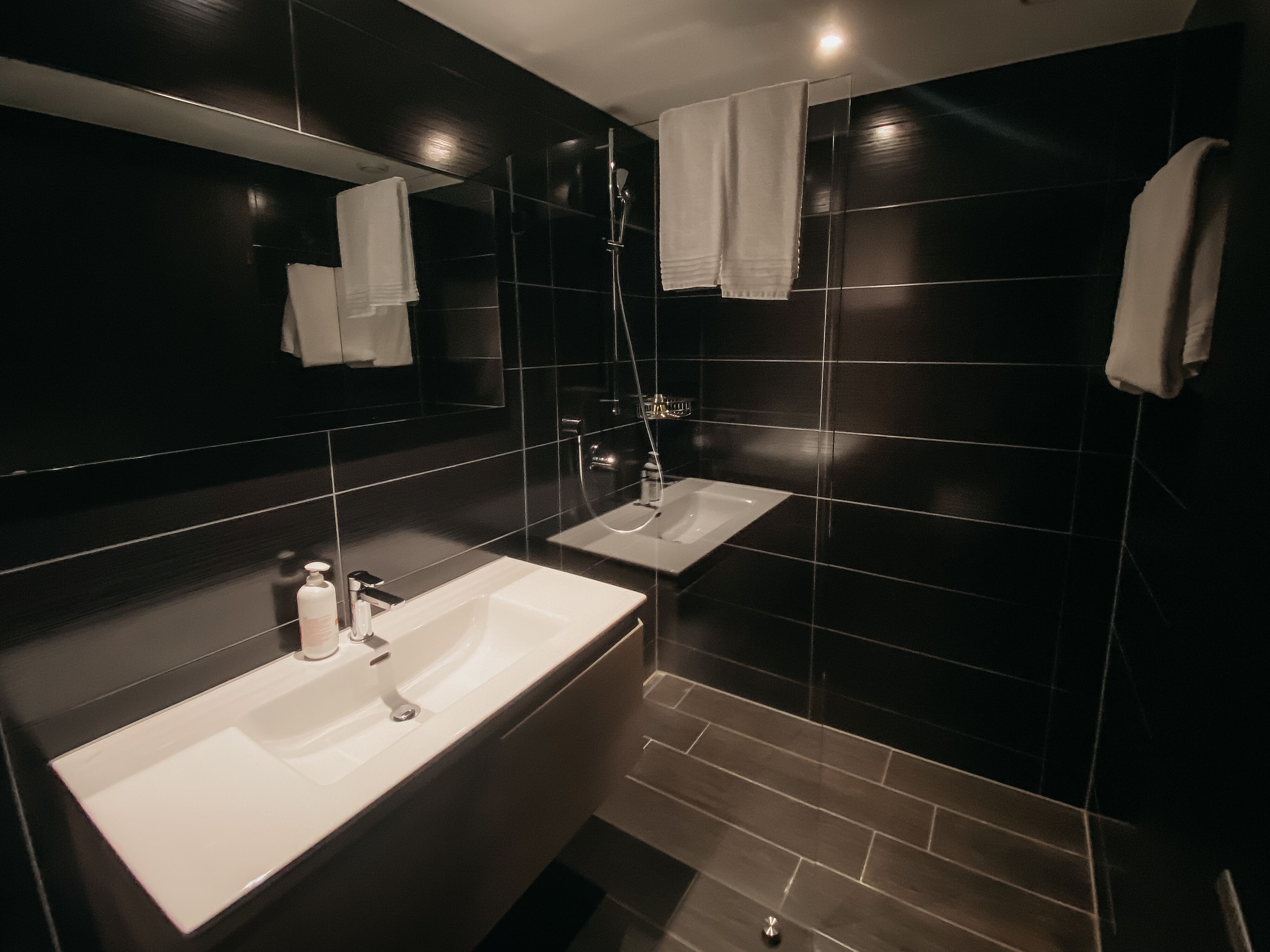Ein modernes Badezimmer mit dunklen Fliesen, zwei Waschbecken, einem Spiegel und einer Dusche.