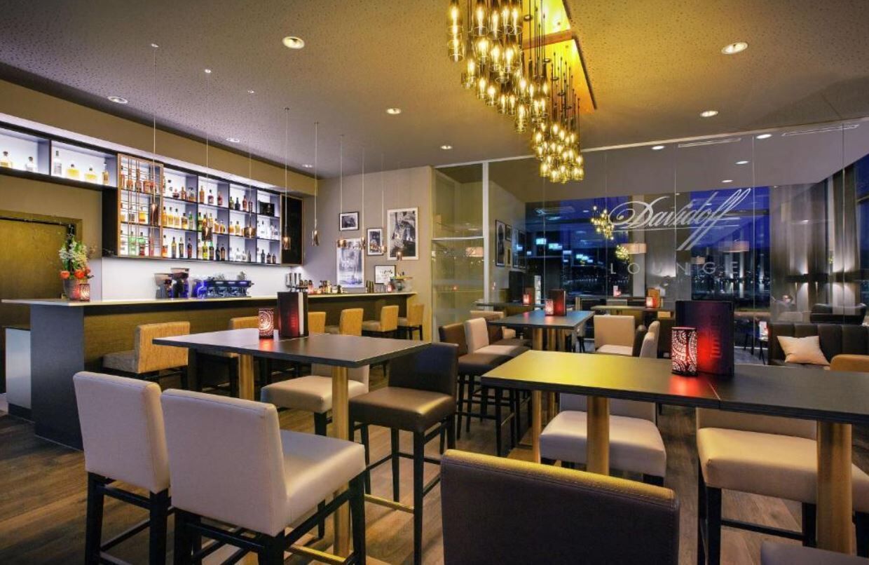 Un bar moderne doté d’un large choix de bouteilles d’alcool, de différents types de sièges et offrant une vue sur la ville grâce à de grandes fenêtres.
