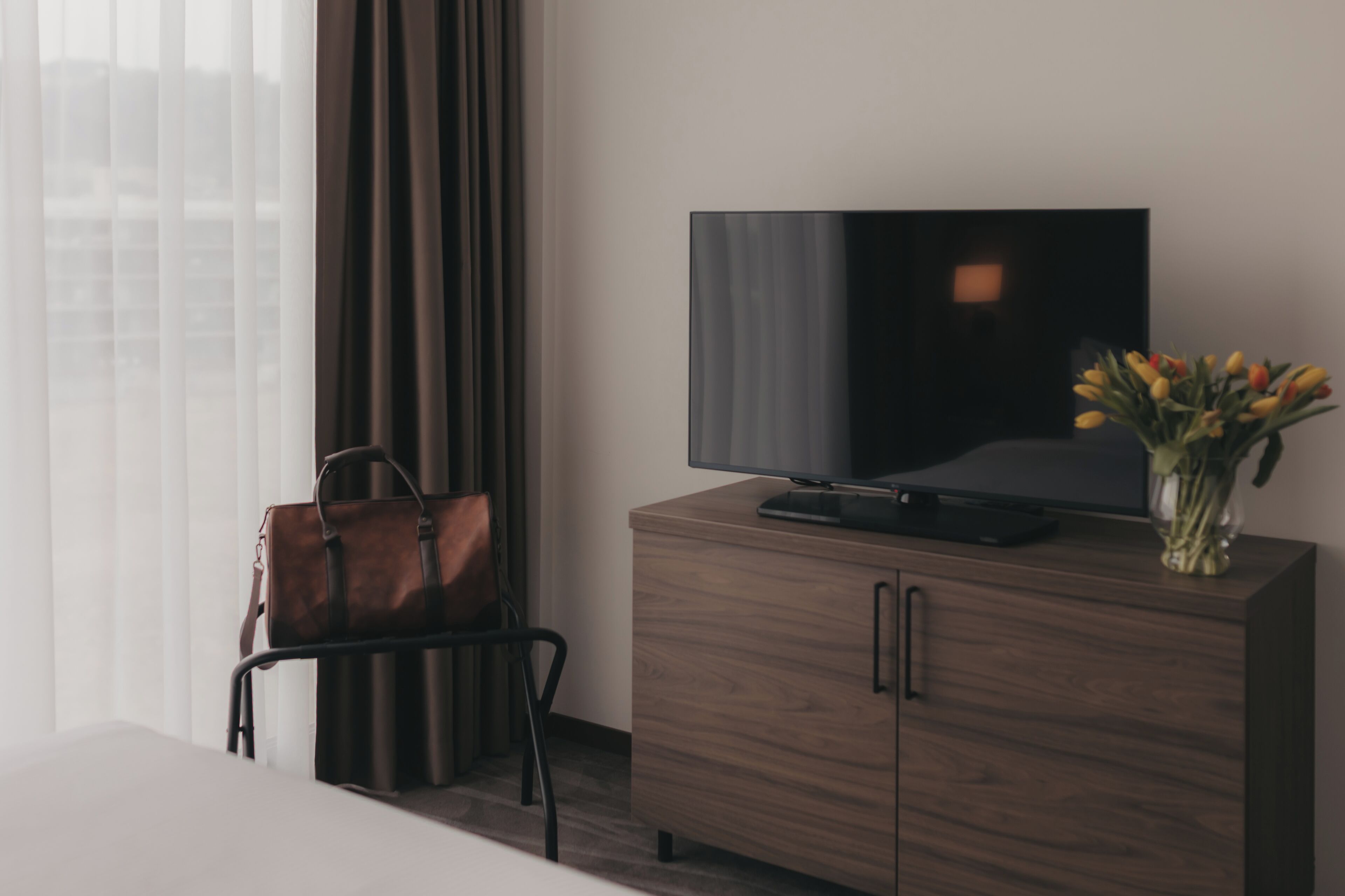 Une chambre d’hôtel moderne avec un meuble en bois, une télévision, un sac en cuir et un vase rempli de fleurs.