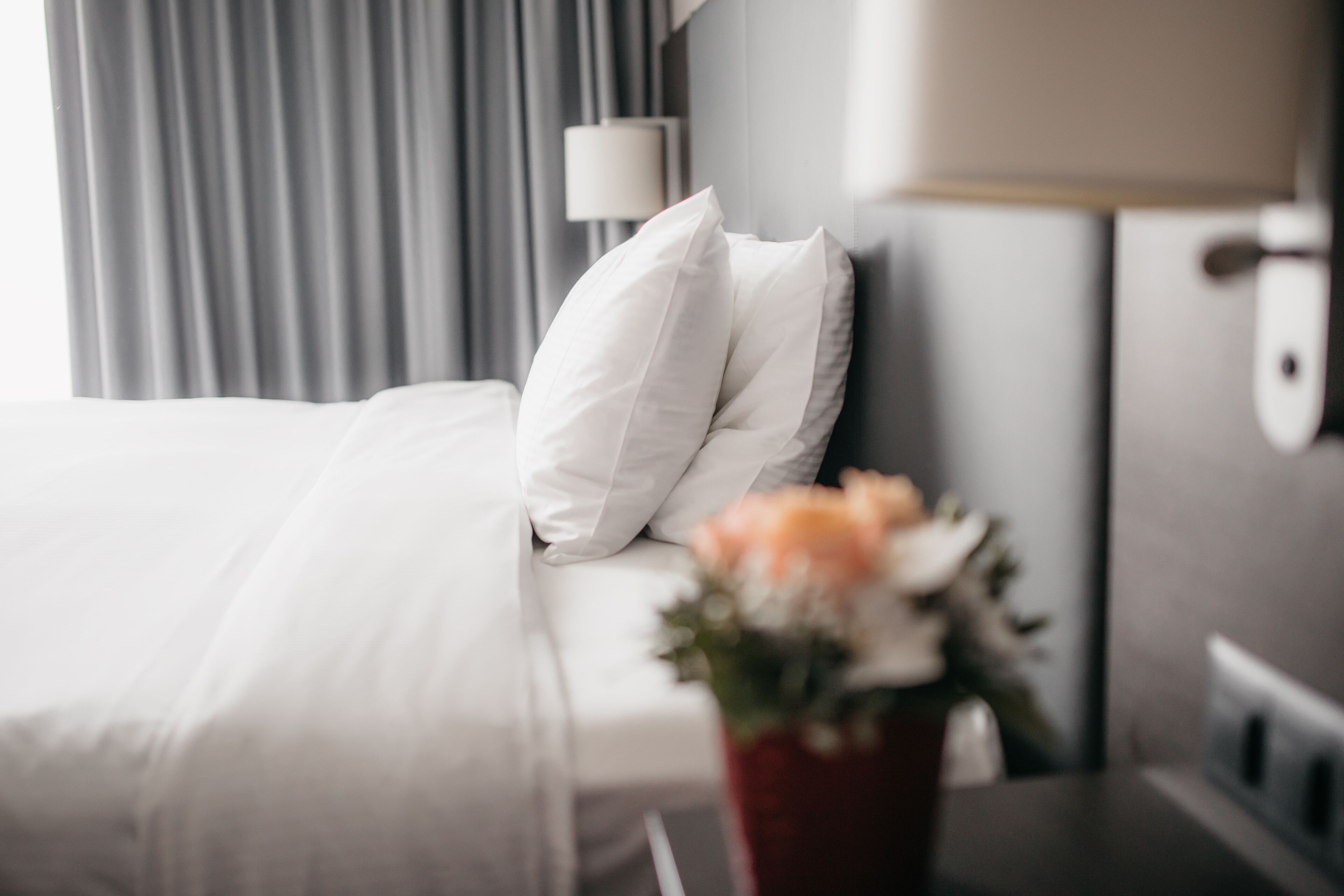 A hotel room with a bed, white pillows, a bedside table with a vase of flowers, and a lamp.