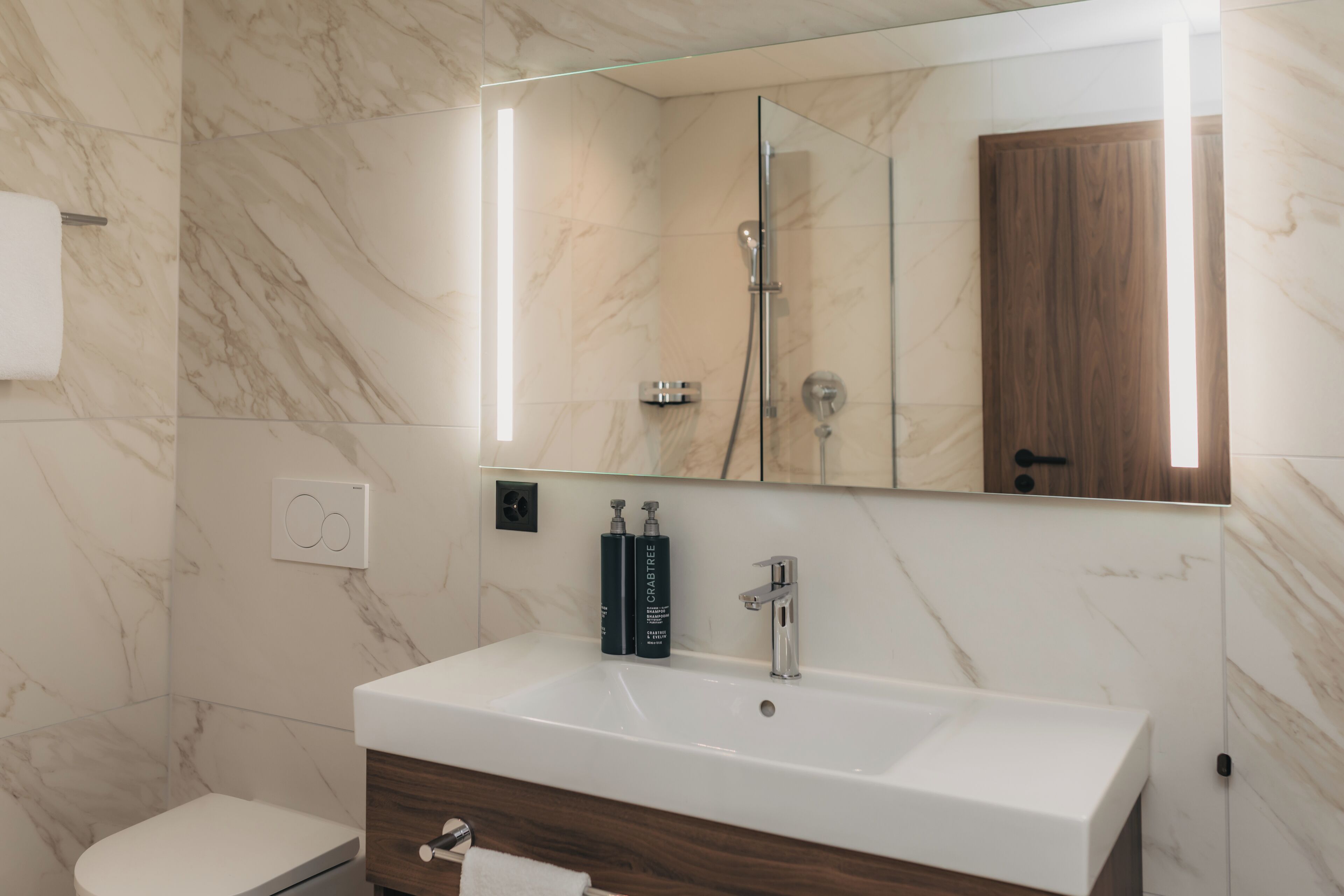 Un baño moderno con pared de mármol, lavamanos blanco, tocador de madera y cabina de ducha con mampara de vidrio.