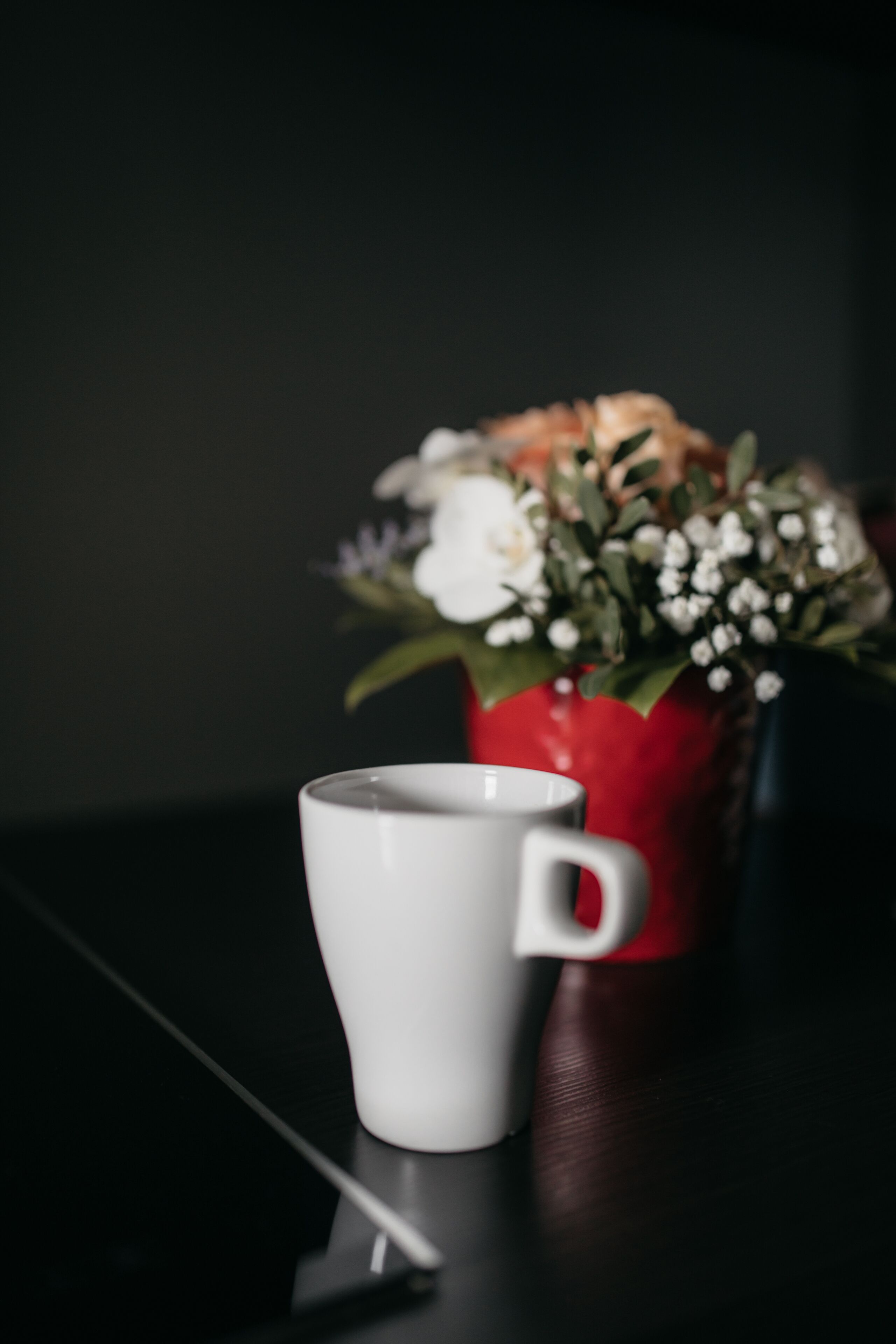 A white mug on a reflective surface with a red vase containing white flowers in the background.