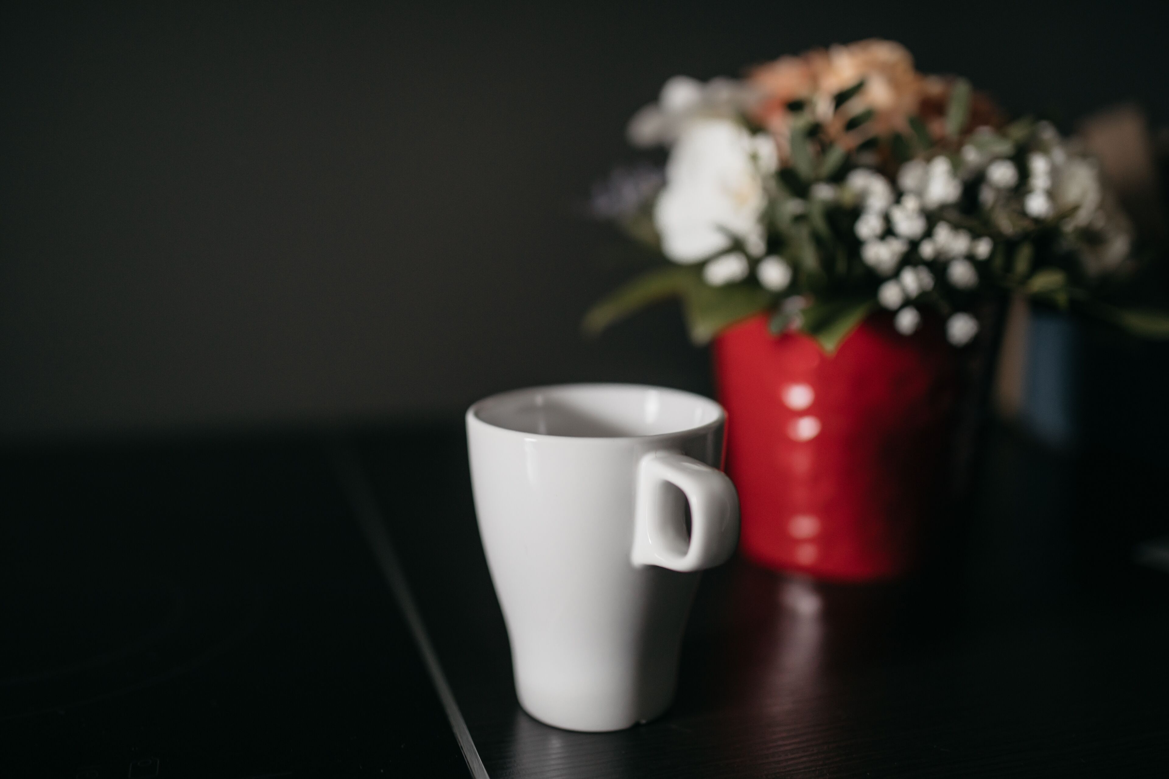 Una tazza bianca su una superficie scura, con un vaso rosso e fiori sullo sfondo.