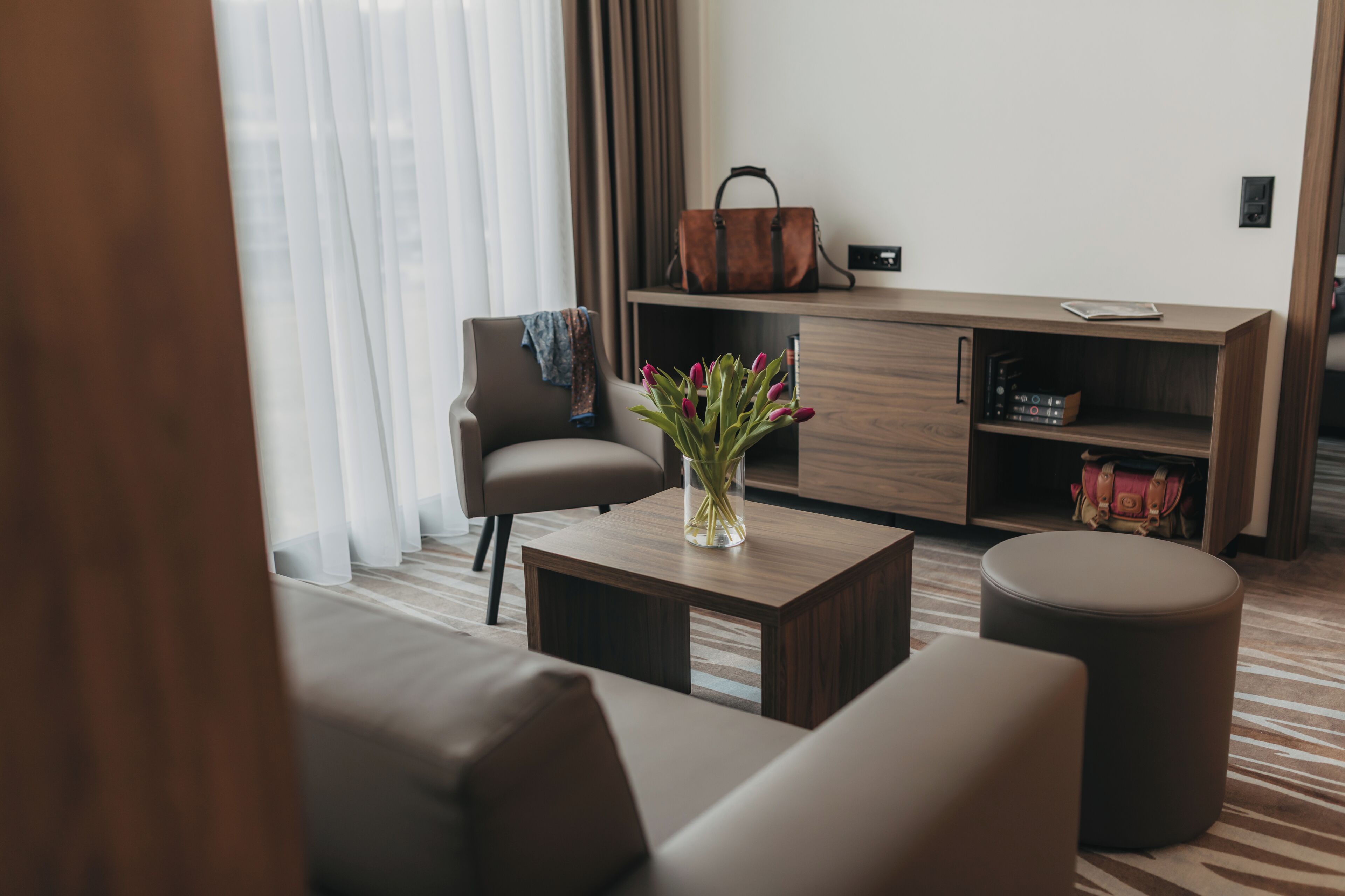 A modern hotel room with a sofa, a coffee table with a vase of flowers, a wooden sideboard, and a leather bag.