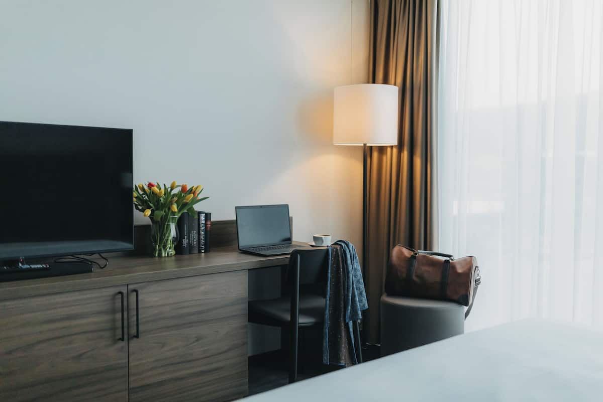 A hotel room with a wooden desk, a laptop, a television, a lamp, a chair, and a suitcase.