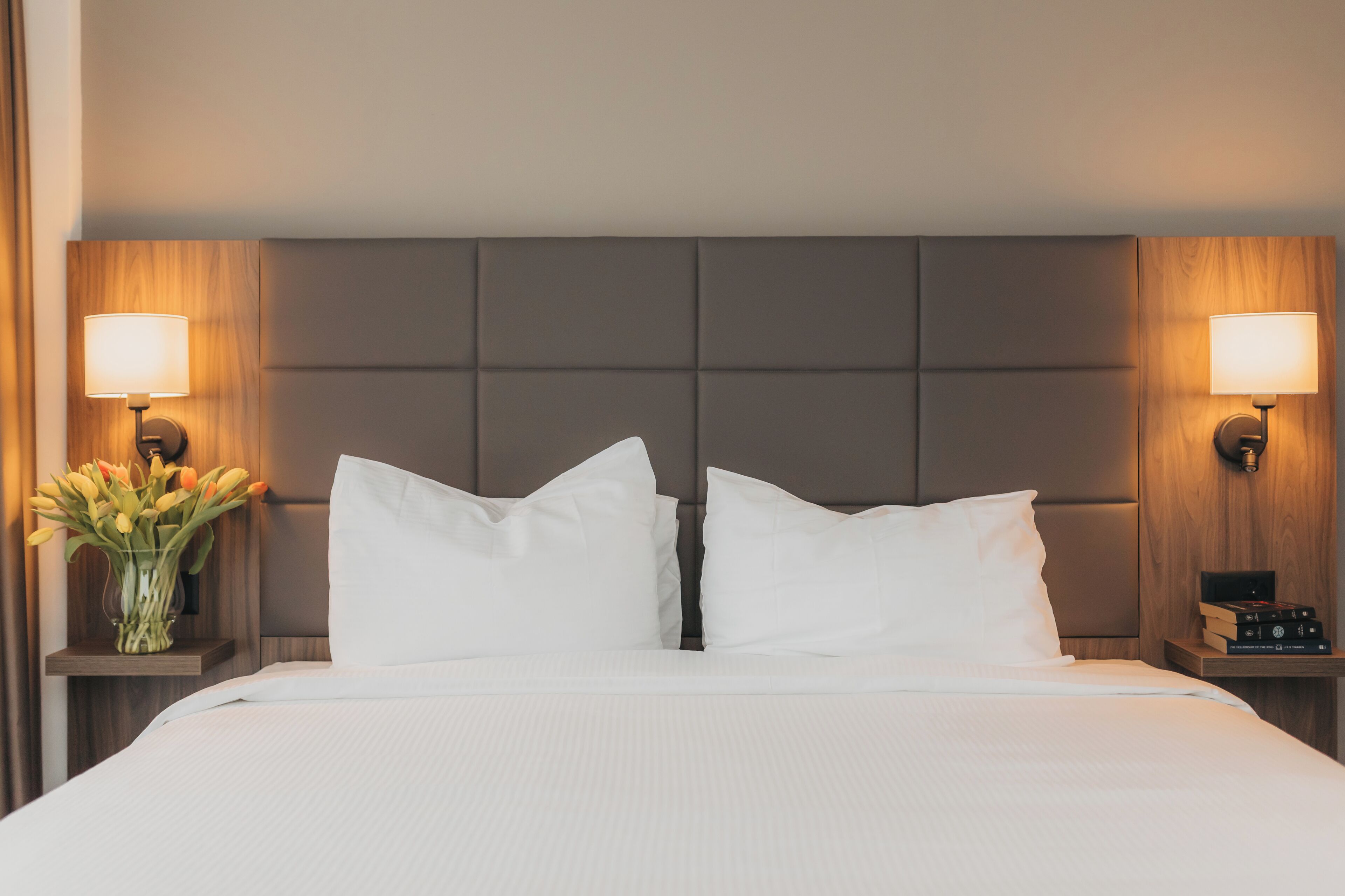 Habitación de hotel con una cama grande, dos lámparas de noche, un florero con flores y una mesita de noche con libros.