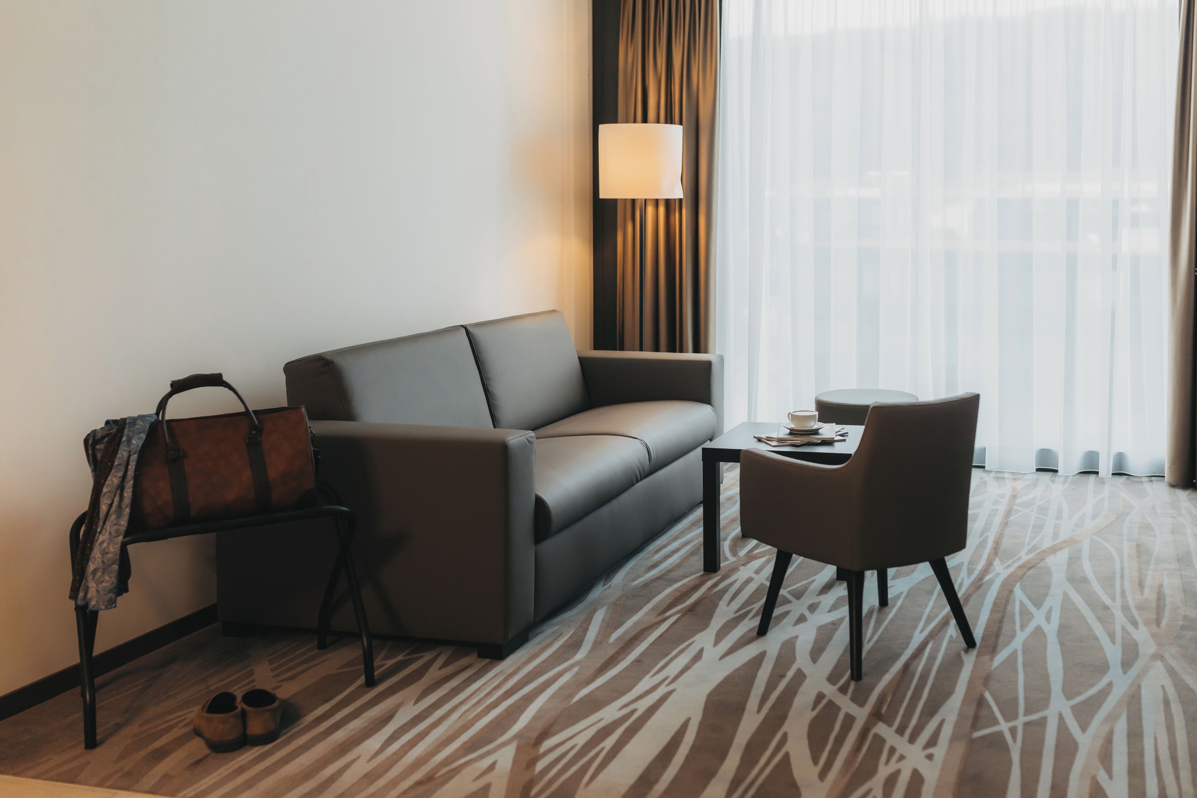 A modern hotel room with a grey sofa, a small table, a chair, and a lamp.